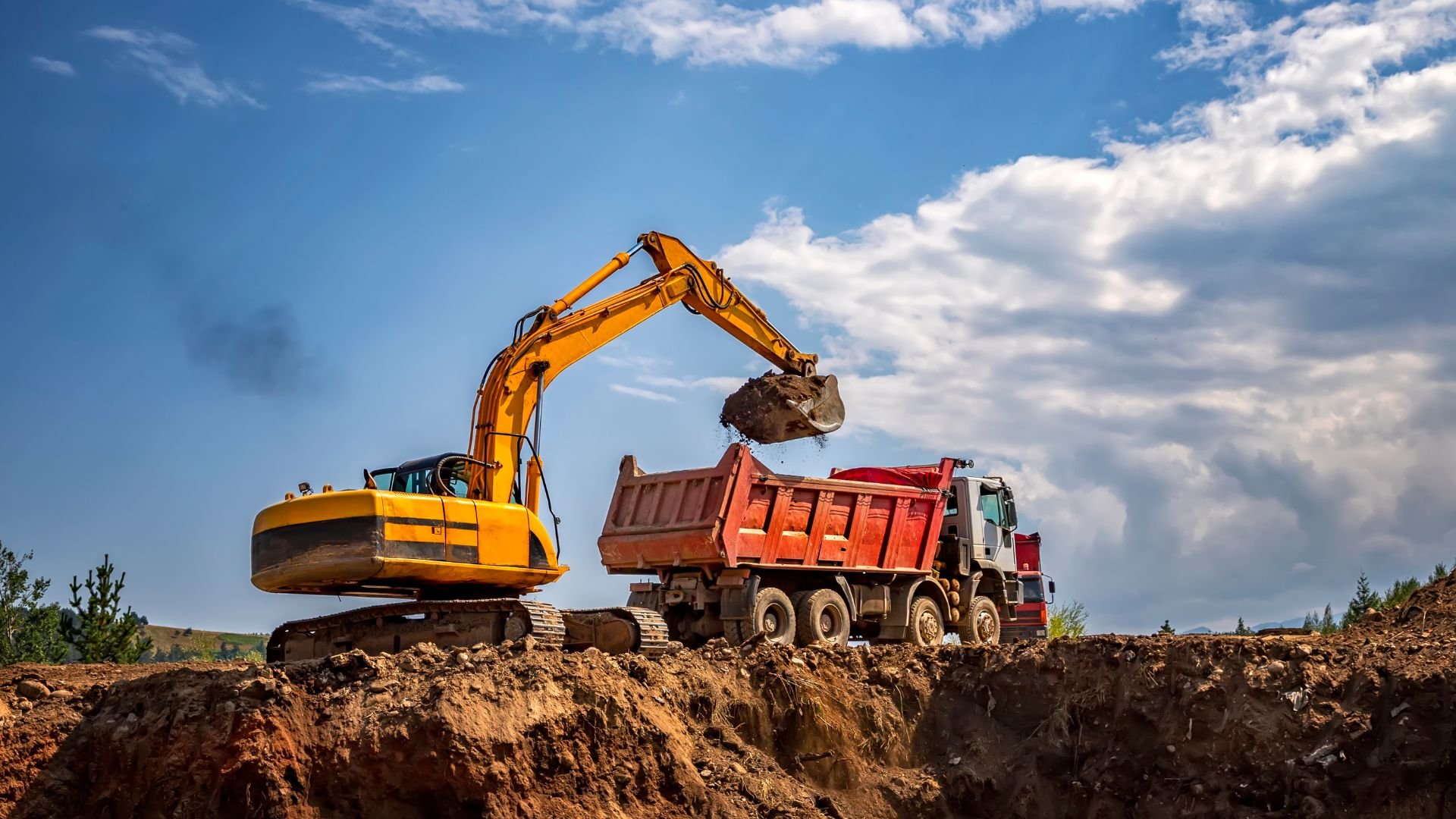 Yellow excavator loading dirt into red dump truck at construction site